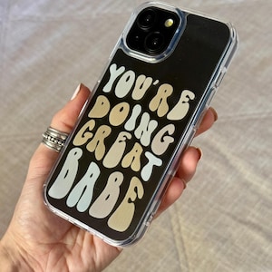 May include: A clear smartphone case featuring a black background with the text YOU'RE DOING GREAT BABE printed in a wavy, multi-colored font consisting of cream, tan, beige, and light blue letters, with a camera cutout at the top corner.