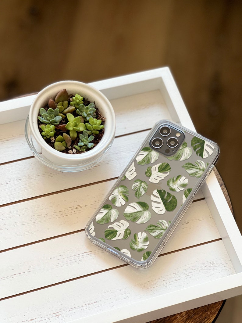 May include: A clear phone case with a green and white tropical leaf pattern. The case is on a white tray with a small potted succulent plant.