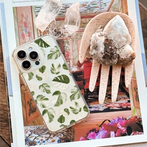 Peut inclure: Une coque de t&eacute;l&eacute;phone transparente avec un motif de feuilles tropicales vertes et blanches. La coque est pos&eacute;e sur un magazine avec le texte "ARCHITECTURAL DIGEST" sur la couverture.