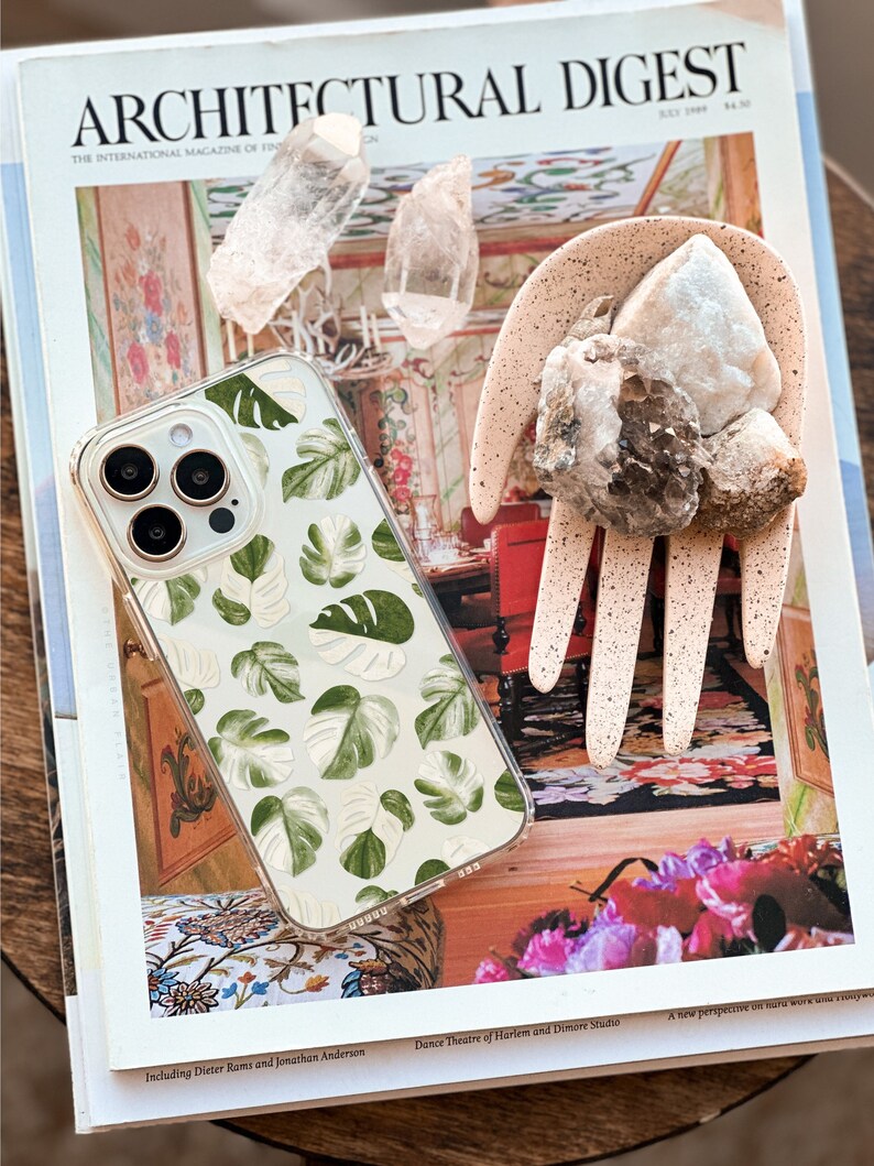 Peut inclure: Une coque de t&eacute;l&eacute;phone transparente avec un motif de feuilles tropicales vertes et blanches. La coque est pos&eacute;e sur un magazine avec le texte "ARCHITECTURAL DIGEST" sur la couverture.