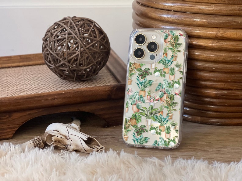 May include: A clear phone case with a floral pattern of potted plants in shades of green, pink, and white. The phone case is designed for an iPhone with a gold camera lens ring.