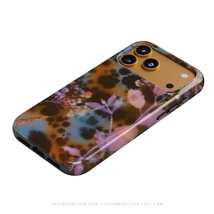 Puede incluir: Una funda para smartphone con un patr&oacute;n oscuro y moteado en tonos marrones, azules y morados con un estampado bot&aacute;nico de hojas y flores. El m&oacute;dulo de la c&aacute;mara es de color dorado con tres aberturas circulares y un flash peque&ntilde;o.