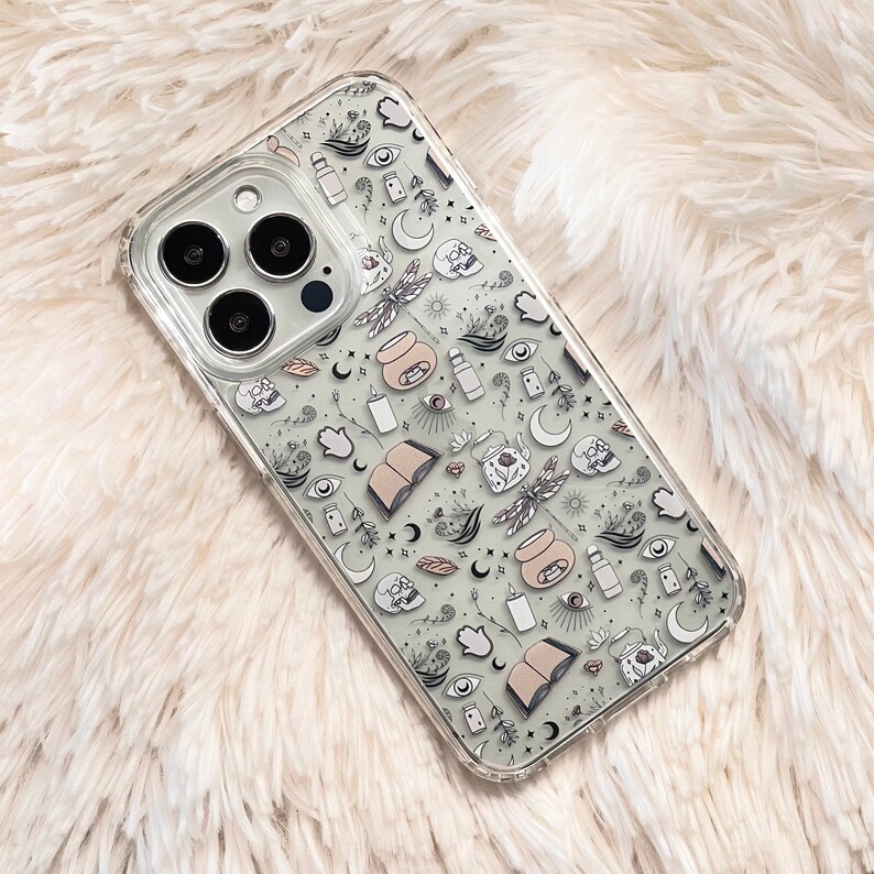May include: A clear phone case with a white background and a black and white pattern of various mystical symbols, including moons, stars, eyes, hands, and a dragonfly.