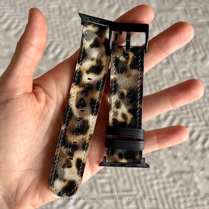 Puede incluir: Una correa de reloj con un estampado de leopardo en tonos marrón, negro y beige. La correa incluye herrajes negros, una hebilla negra y dos trabillas negras. El texto THEURBANFLAIR.COM está impreso en letras blancas pequeñas abajo.