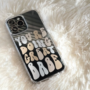 May include: A clear phone case with a black background and a gold and white sticker that says "You're doing great babe".
