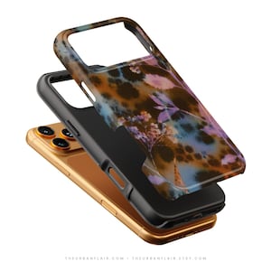 Puede incluir: Un tel&eacute;fono inteligente dorado colocado bajo una funda protectora negra con un dise&ntilde;o floral decorativo en tonos marr&oacute;n, azul y morado. La funda tiene recortes para la c&aacute;mara y los botones laterales, mostrando un dise&ntilde;o en capas.