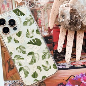 Peut inclure: Une coque de t&eacute;l&eacute;phone transparente avec un fond blanc et un motif de feuilles vertes et blanches. La coque est con&ccedil;ue pour un iPhone 14 Pro.