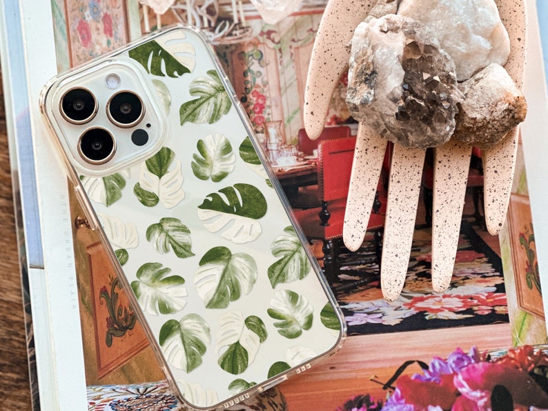 Peut inclure: Une coque de t&eacute;l&eacute;phone transparente avec un fond blanc et un motif de feuilles vertes et blanches. La coque est con&ccedil;ue pour un iPhone 14 Pro.