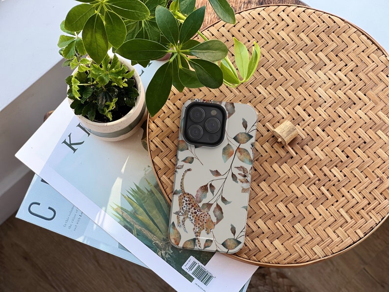 Pu&ograve; includere: Una custodia bianca per iPhone con un design ad acquerello di foglie e una stampa leopardata. La custodia &egrave; su una superficie di bamb&ugrave; intrecciato.