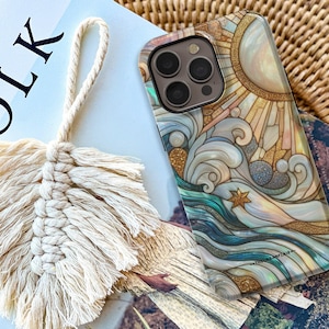 May include: A phone case featuring a stained-glass style design of a sun, swirling ocean waves, and a star, with gold glitter accents. Beside it is a cream-coloured macrame feather keychain with a braided loop, both resting on a book cover.