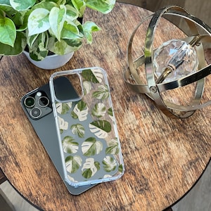 Peut inclure: Une coque de t&eacute;l&eacute;phone transparente avec un motif de feuilles tropicales vertes et blanches. La coque est pos&eacute;e sur une table en bois avec un orbe m&eacute;tallique d&eacute;coratif.