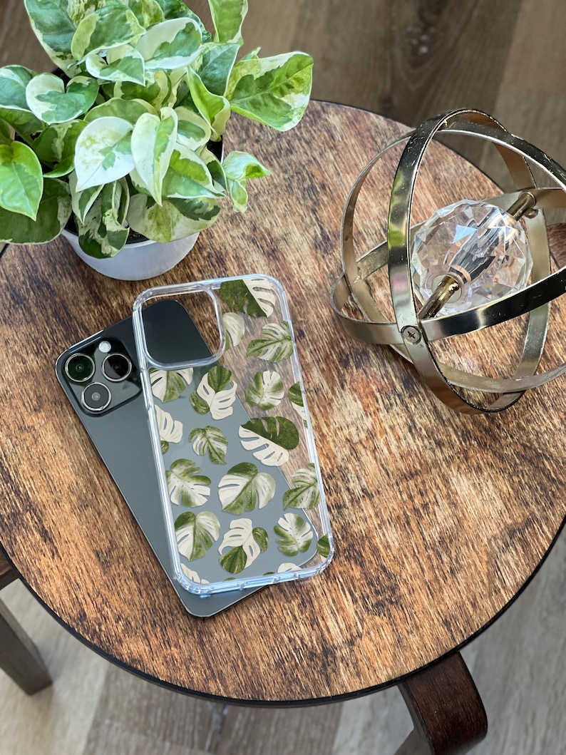 Peut inclure: Une coque de t&eacute;l&eacute;phone transparente avec un motif de feuilles tropicales vertes et blanches. La coque est pos&eacute;e sur une table en bois avec un orbe m&eacute;tallique d&eacute;coratif.