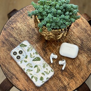 Peut inclure: Une coque de t&eacute;l&eacute;phone transparente avec un motif de feuilles tropicales vertes et blanches, une paire d'&eacute;couteurs sans fil blancs et un pot de succulentes dor&eacute; sur une table en bois.
