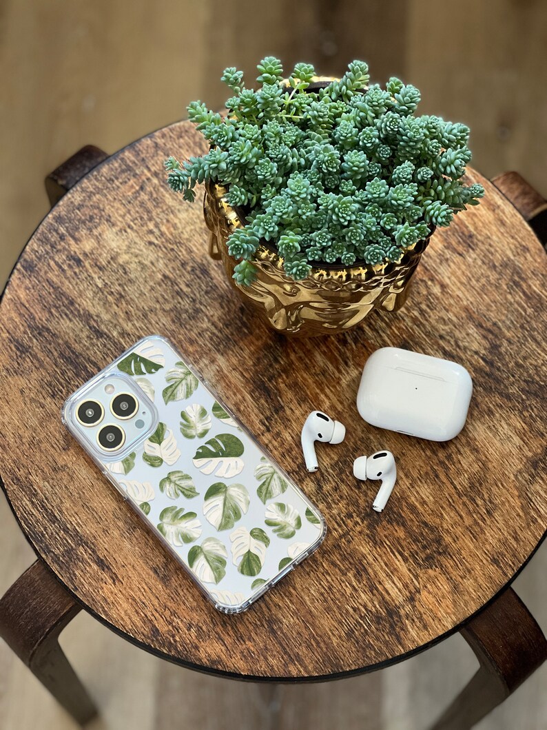 Peut inclure: Une coque de t&eacute;l&eacute;phone transparente avec un motif de feuilles tropicales vertes et blanches, une paire d'&eacute;couteurs sans fil blancs et un pot de succulentes dor&eacute; sur une table en bois.