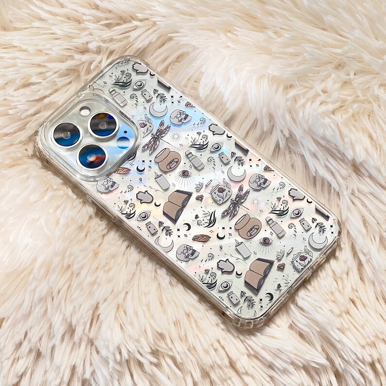 May include: A clear phone case with a holographic design featuring various symbols and illustrations, including a dragonfly, an eye, a hand, and a book. The case is designed for an iPhone.