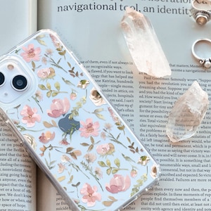 Puede incluir: Funda transparente para teléfono con un estampado floral de flores rosas y melocotón, hojas verdes y pequeñas flores blancas. La funda está sobre una página de libro con anillos y cristales.