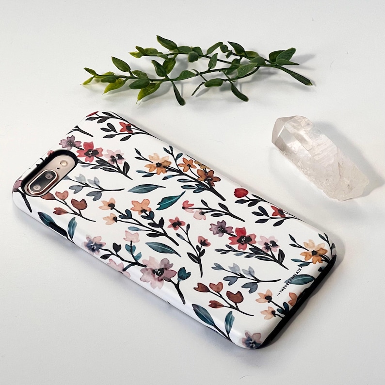 May include: A white phone case with a floral pattern featuring watercolor-style flowers in shades of pink, red, orange, and yellow. The flowers are arranged on a white background with dark gray stems and leaves.