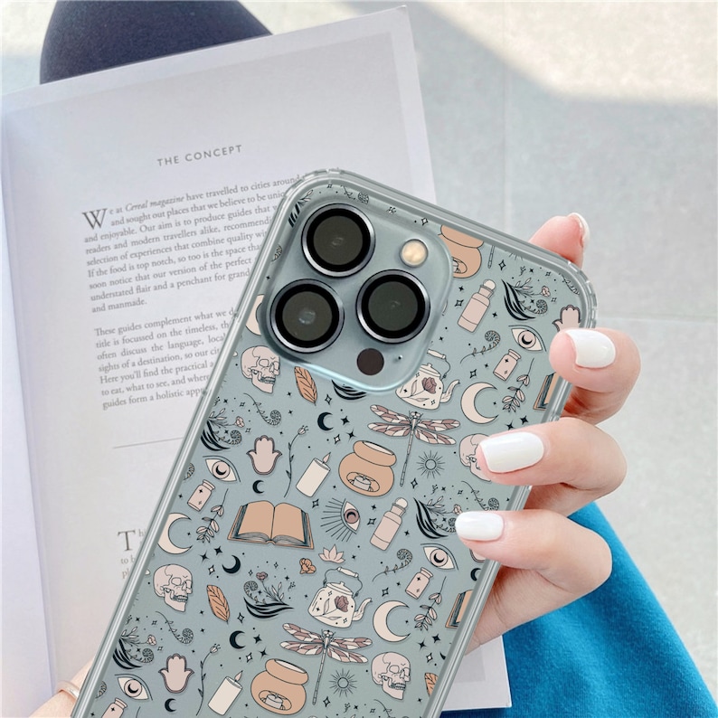 May include: A clear phone case with a blue and white design featuring a variety of illustrations including skulls, moons, stars, teapots, candles, and dragonflies.