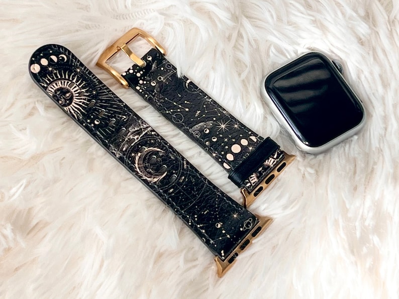 Peut inclure: Un bracelet de montre en cuir noir avec une boucle dor&eacute;e et un imprim&eacute; c&eacute;leste repr&eacute;sentant des lunes, des &eacute;toiles et un soleil. Le bracelet est con&ccedil;u pour une Apple Watch.