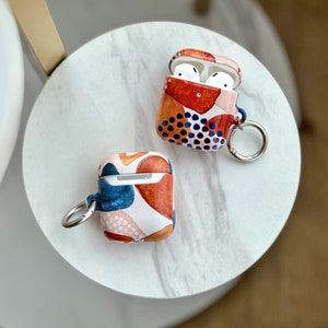 Puede incluir: Dos estuches para AirPod con patrones geom&eacute;tricos abstractos en tonos rojos, azules y blancos. Ambos estuches tienen un anillo de metal plateado adjunto para un llavero.