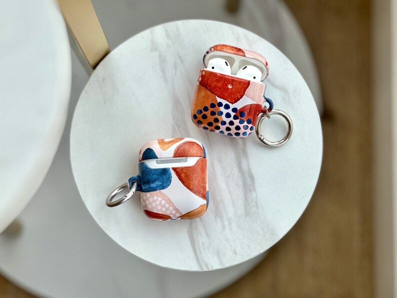 Puede incluir: Dos estuches para AirPod con patrones geom&eacute;tricos abstractos en tonos rojos, azules y blancos. Ambos estuches tienen un anillo de metal plateado adjunto para un llavero.