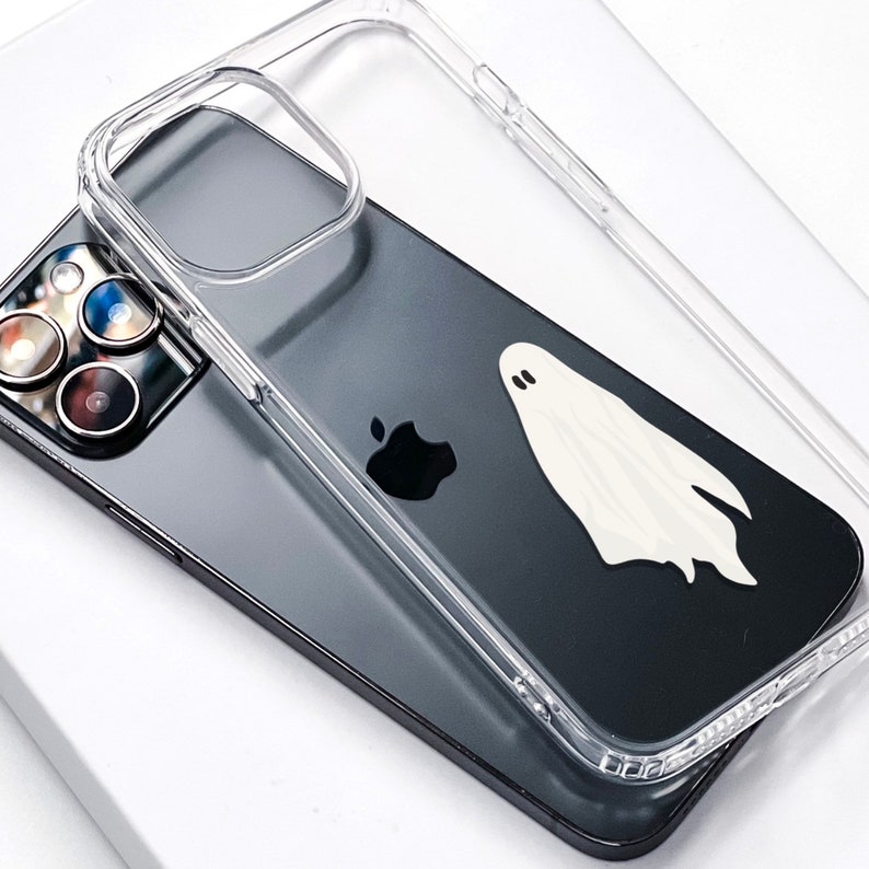 May include: A clear phone case with a ghost design on a black iPhone with a camera lens.