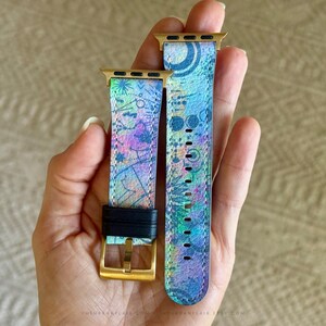 K&ouml;nnte beinhalten: Ein zweiteiliges Uhrenarmband mit einem lebendigen, mehrfarbigen abstrakten Muster in Blau-, Lila- und Gr&uuml;nt&ouml;nen. Das Armband verf&uuml;gt &uuml;ber goldfarbene Metallanschl&uuml;sse und eine goldfarbene Schlie&szlig;e mit einer schwarzen Lederschlaufe.