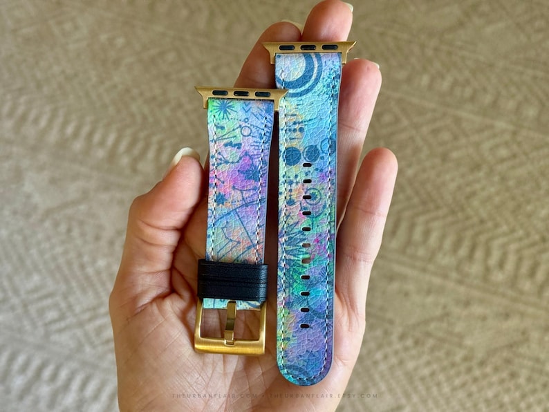 K&ouml;nnte beinhalten: Ein zweiteiliges Uhrenarmband mit einem lebendigen, mehrfarbigen abstrakten Muster in Blau-, Lila- und Gr&uuml;nt&ouml;nen. Das Armband verf&uuml;gt &uuml;ber goldfarbene Metallanschl&uuml;sse und eine goldfarbene Schlie&szlig;e mit einer schwarzen Lederschlaufe.