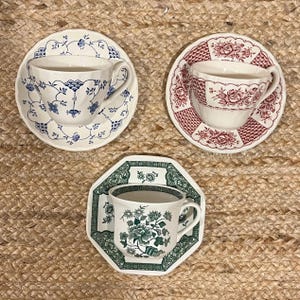 May include: Three teacups and saucers with floral patterns. The teacups are white with blue, red, and green floral designs. The saucers are white with matching floral designs.