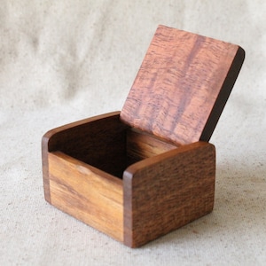 May include: A small wooden box with a hinged lid, made of dark brown wood with a natural wood grain. The lid is open, revealing the empty interior of the box.