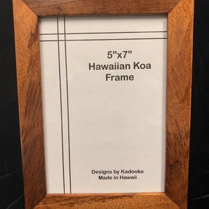 May include: A 5" x 7" wood picture frame made of Hawaiian Koa wood. The frame has a dark brown color and a natural wood grain. The frame is made in Hawaii and has a label that says "Designs by Kadooka Made in Hawaii".