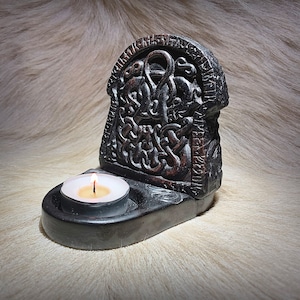 May include: Black candle holder with a carved design of a knotwork pattern. The holder has a small, round indentation for a tea light candle.