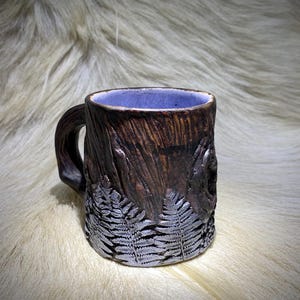 May include: A handcrafted ceramic mug with a brown, textured exterior resembling tree bark. The mug features a fern leaf design and a purple interior. The handle is also textured to match the body. A unique, nature-inspired piece.