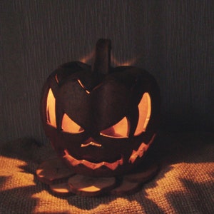 Può includere: Una zucca intagliata con un'espressione minacciosa brilla di una luce arancione da una candela all'interno. La zucca è appoggiata su un pezzo di legno su una superficie di juta.