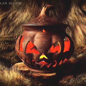 Può includere: Una lanterna di ceramica marrone a forma di zucca con un viso di zucca intagliato. La lanterna è illuminata dall'interno, creando un bagliore caldo.