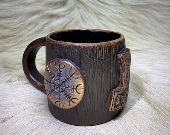 May include: A brown ceramic mug with a wood grain design. The mug has a handle and features a gold-colored Viking symbol and a gold-colored compass.
