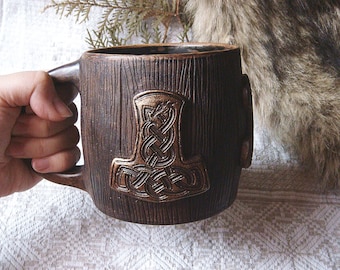 May include: A brown ceramic mug with a wood grain design and a raised, gold-colored Mjolnir symbol. The mug has a handle on the side.