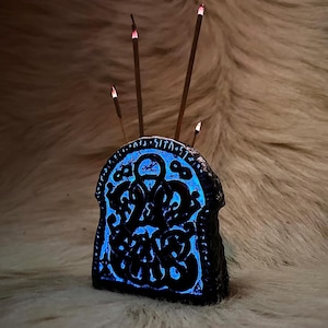 May include: A black ceramic incense holder with a blue glow and intricate carvings. Four incense sticks are placed in the holder.