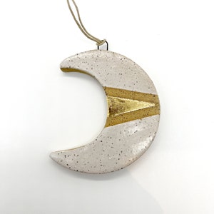 May include: A ceramic crescent moon ornament with a speckled off-white glaze. A gold-colored band with a triangle design accents the center. A tan string is attached for hanging. The ornament is approximately 4 inches wide.