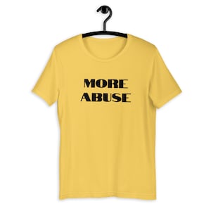 Può includere: Una maglietta gialla a maniche corte con la scritta "MORE ABUSE" stampata in nero.