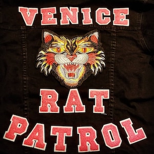 May include: A black denim jacket with pink embroidered patches that say "Venice", "Rat", and "Patrol". A fierce orange and yellow embroidered cat face is centered above the words "Rat".