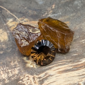 May include: A faceted, round, dark brown gemstone with a brilliant cut, displayed with two rough, amber-colored mineral specimens. The gemstones are set against a textured, neutral-toned surface.