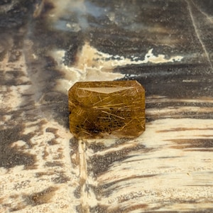 Puede incluir: Una gema de color marrón dorado con forma rectangular. La piedra presenta una red de finas inclusiones en forma de cabello, creando un patrón único. La gema se encuentra sobre un fondo moteado de tonos neutros.