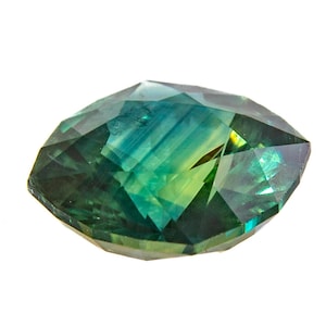 May include: A faceted gemstone with a marquise cut. The gem displays a blend of green and blue hues, with a hint of yellow. The facets create a play of light and shadow, enhancing its brilliance. The gemstone is set against a white background.