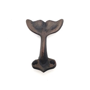 May include: A cast iron wall hook shaped like a whale tail. The hook is a dark brown colour and has a rustic finish.