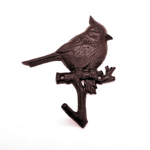 May include: A dark brown cast iron wall hook shaped like a bird on a branch. The hook has a detailed bird design and a textured surface. The hook is mounted on a white background. The hook is suitable for hanging items.