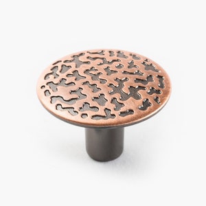 Puede incluir: Un pomo de armario redondo de color cobre con un diseño negro intrincado y estampado. El pomo tiene un vástago corto negro.