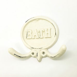 以下が含まれることがあります： 白い鋳鉄製のダブルフック。丸い看板に黒い文字で「BATH」と書かれています。