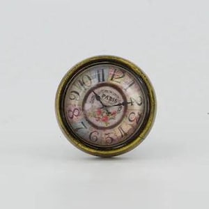 Puede incluir: Un pomo de puerta redondo en forma de reloj, de color dorado, con una esfera blanca y números romanos negros. La esfera del reloj tiene un diseño floral y la palabra "París" impresa en ella.