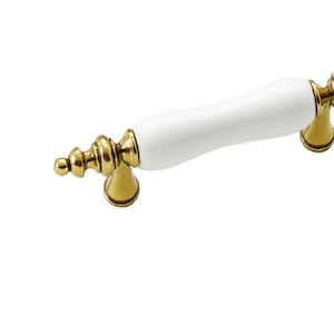 Peut inclure: Poignée de cabinet en céramique blanche avec des accents en métal doré. La poignée a une forme rectangulaire arrondie et mesure environ 10 cm de long.